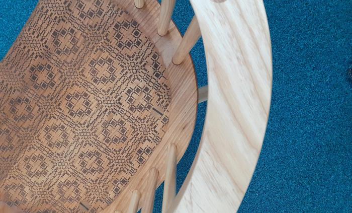 Aerial view looking down on to a wooden chair. The curved arm of the chair can be seen above a number of dowel features leading down to a highly patte